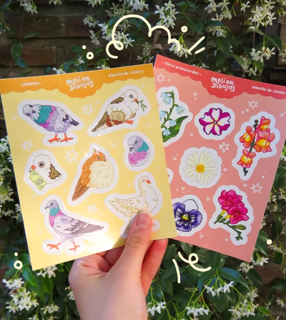 Foto de dos planchas de stickers ilustrados. Una, amarilla, es sobre diferentes palomas. La otra, rosa, tiene diferentes flores primaverales.