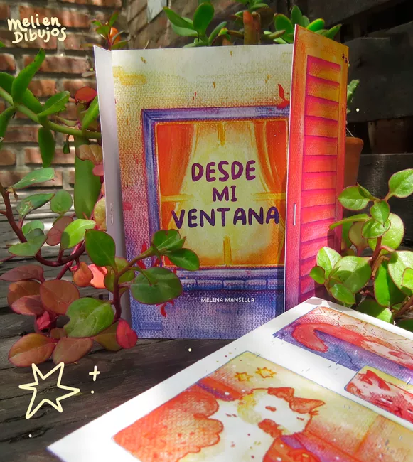 Foto de la portada del fanzine Desde mi ventana, que muestra una ventana luminosa, de cortinas naranjas y persianas rosas y violetas. Frente a este, recostado, se ve parcialmente una página interior del fanzine sobre un gato naranja y blanco.