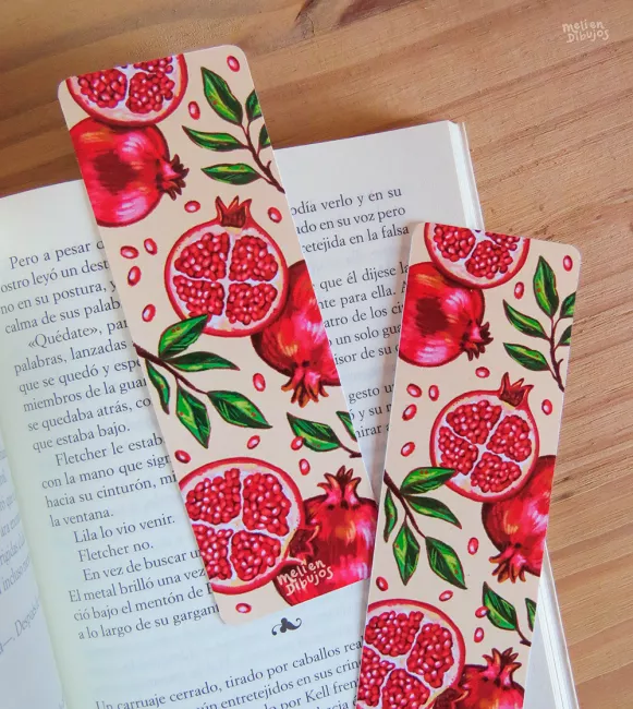 Dos marcapáginas con un patrón de granadas, enteras y cortadas, y en colores rojos, verdes y beige, sobre un libro abierto.