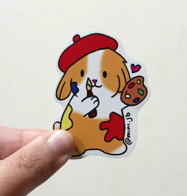 Sticker de un conejo caricaturizada con boina roja, sosteniendo un pincel y una paleta de colores. Tiene manchas de pintura en su cuerpo y una pequeña figura de corazón rosa al lado. En el borde se lee la firma “@mini.jb”.