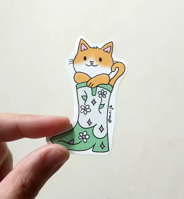 Sticker de un gato anaranjado y blanco dentro de una bota verde decorada con flores y rombos violetas. El gato asoma con expresión alegre y la firma “@mini.jb” aparece al costado.