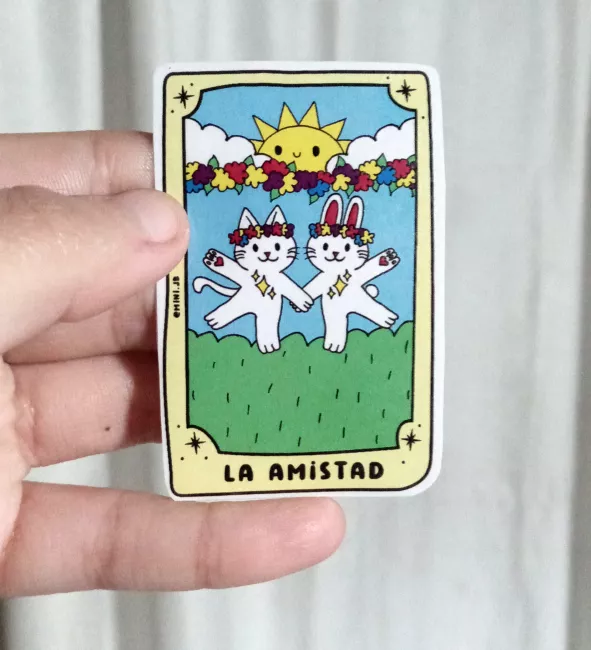 Sticker en forma de carta de tarot titulada “La Amistad”. Se ven dos gatos blancos sonrientes con coronas de flores, tomados de la mano en un campo verde. Detrás hay un arco de flores de colores, nubes y un sol amarillo sonriente en el cielo azul.