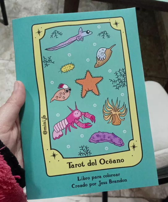 Portada del libro “Tarot del Océano. Libro para colorear”, creado por Jess Brandon. La tapa es de color turquesa y muestra ilustraciones de distintos animales marinos: una estrella de mar, un pez, un caracol, un pulpo pequeño, un crustáceo rosa, un erizo, una anémona y un pepino de mar. La ilustración está enmarcada como si fuera una carta de tarot, con el texto @mini.jb en un costado.