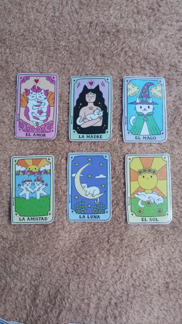 Seis stickers con ilustraciones estilo cartas de tarot. De izquierda a derecha y de arriba hacia abajo: “El Amor” muestra dos gatos abrazados con alas, “La Madre” representa una figura humana con orejas de gato abrazando un gato, “El Mago” es un gato con sombrero y capa de mago; “La Amistad” presenta dos gatos con coronas de flores bailando bajo el sol; “La Luna” muestra un gato durmiendo en una luna creciente rodeada de estrellas; y “El Sol” un gato recostado sobre flores bajo un sol.