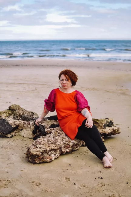 una modelo sentada en un tronco en la playa, con el mar de fondo viste un vestido color coral con mangas hechas con una camisa estampada color fucsia y naranja